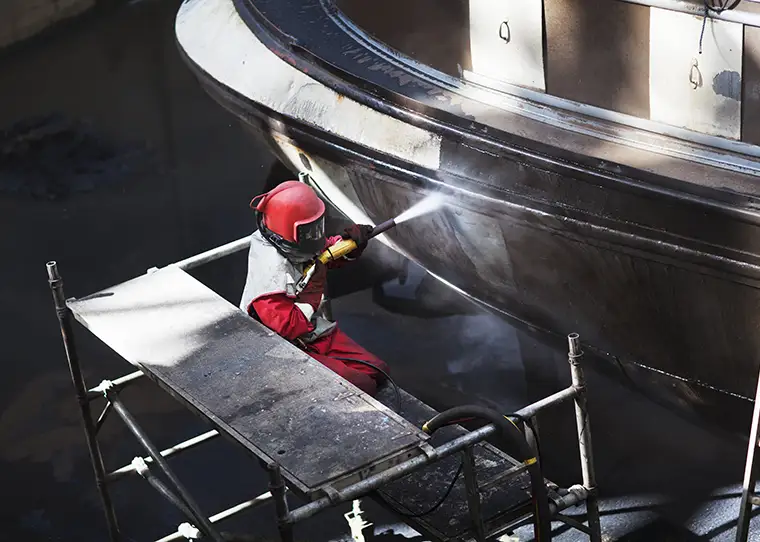 SurfacePrep Worker cleaning hull of boat with blasting hose in shipyard